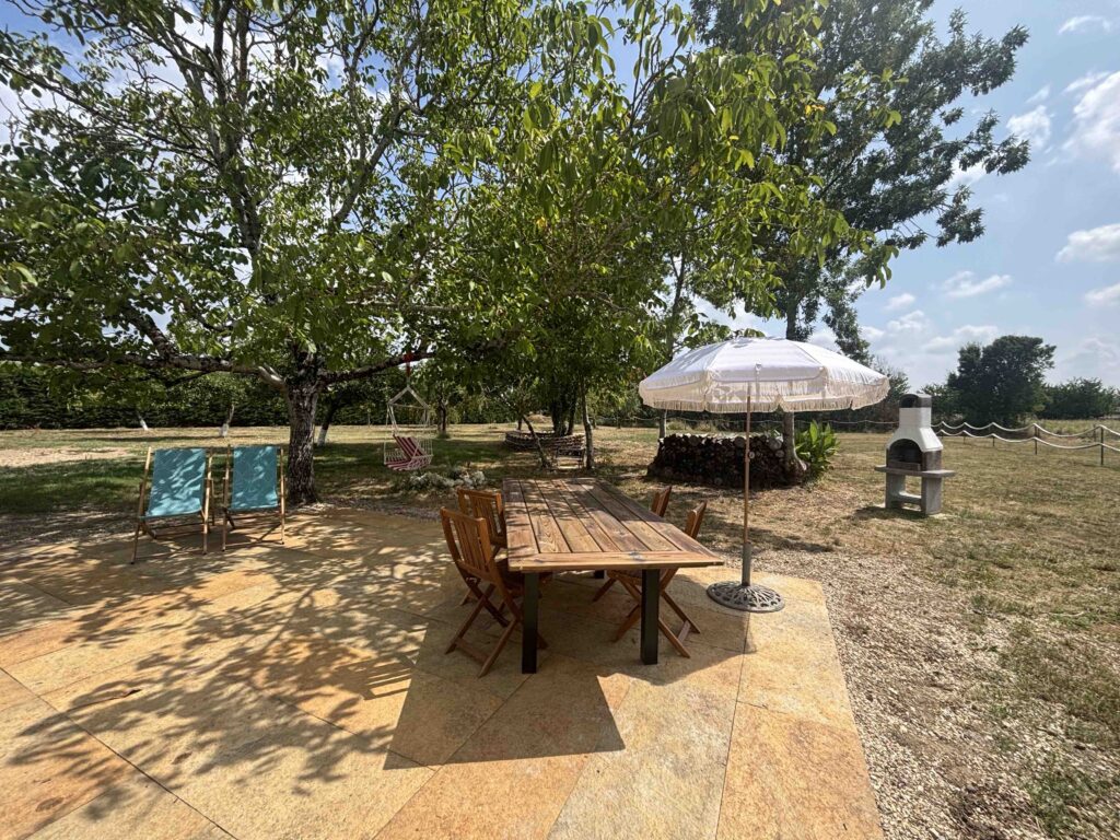 Terrasse en pierre avec table en bois, chaises en acacia, parasol, transat, hamac siège et barbecue à la Maison des Noix, gîte à louer en Dordogne